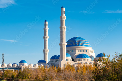 The main central mosque of Astana with blue domes and tall white minarets, located against a clear blue sky, surrounded by trees against an autumn landscape. Kazakhstan