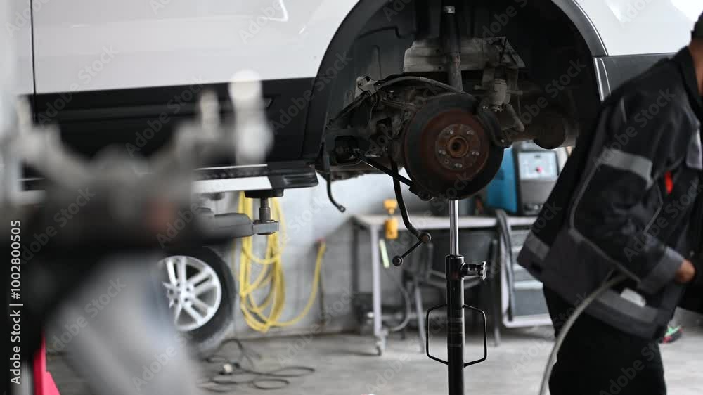 A mechanic in a uniform is working under a white car that is lifted on a jack in a repair shop. He is using a power tool. A mechanic in a uniform is working under a white car that is lifted on a jack in a repair shop. He is using a power tool.