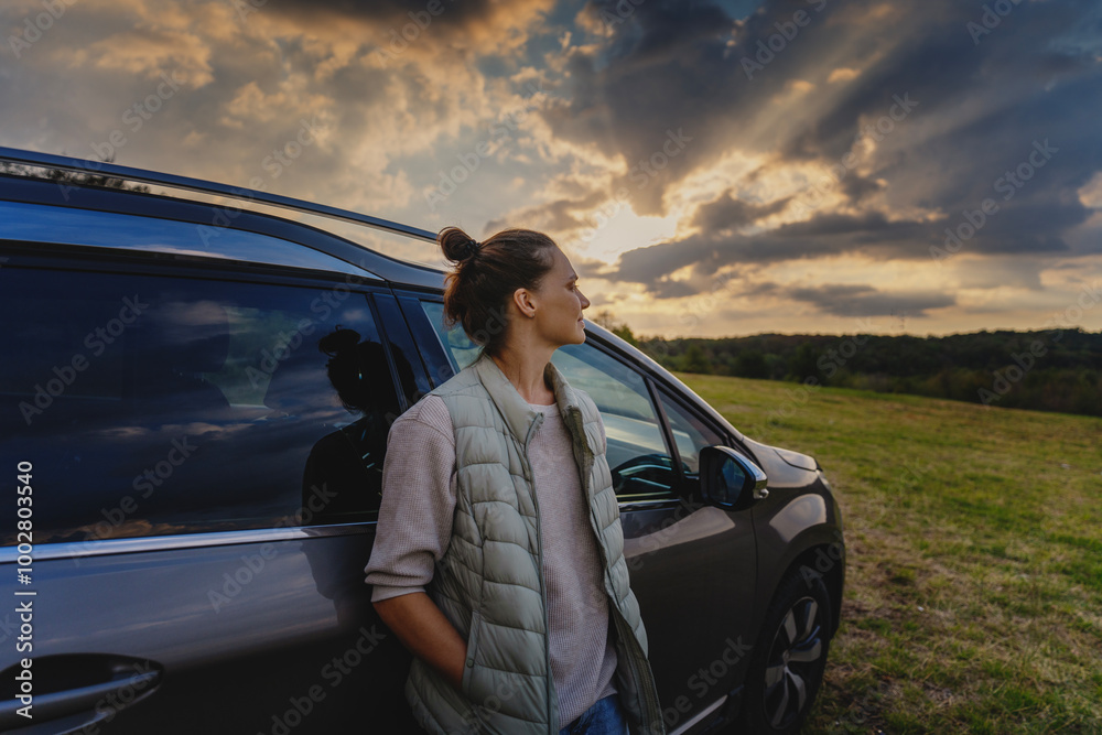 Young female driver standing by car enjoying sunset and nature in field during road trip