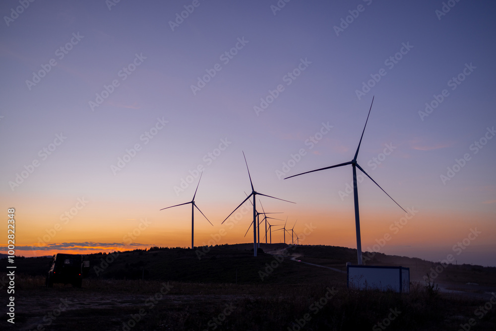 Obraz premium Silhouette of wind turbines against a colorful sunset sky. The gentle fading light highlights the calm beauty of renewable energy in harmony with nature. A peaceful moment at dusk