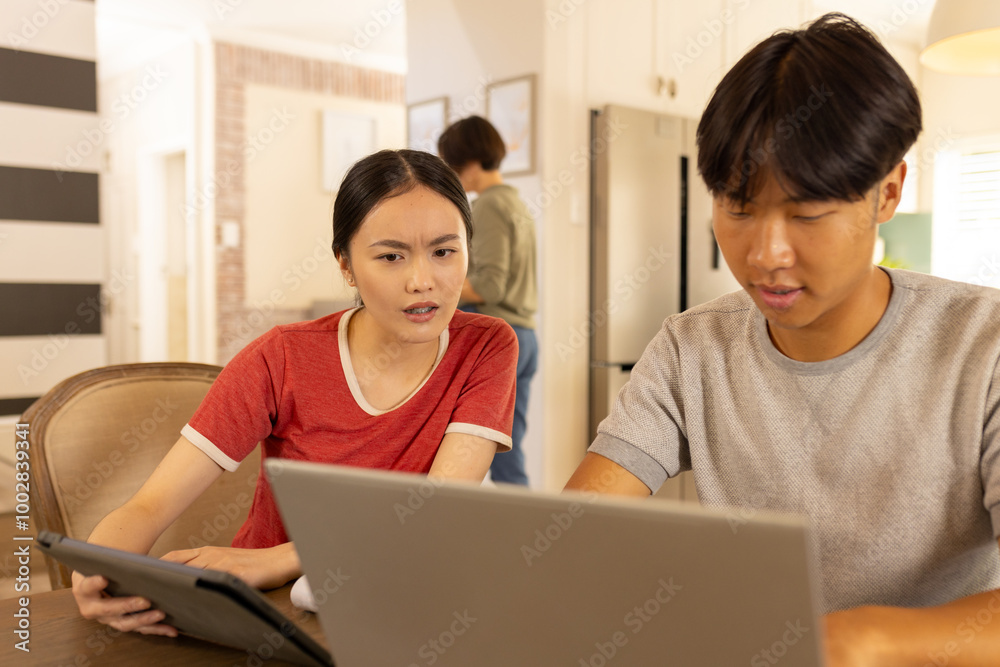 Using laptop and tablet, young asian brother and sister discussing something in kitchen Using laptop and tablet, young asian brother and sister discussing something in kitchen