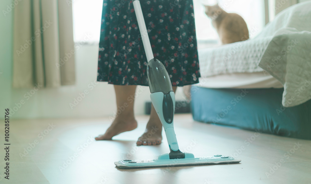 A woman is cleaning the floor with a mop. A cat is sitting on the bed