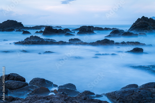 At dusk. Gentle waves lap against smooth, rounded rocks in a tranquil cove. The sky is painted in soft hues of pink and purple, reflecting on the calm water. Toucheng, Yilan, Taiwan.