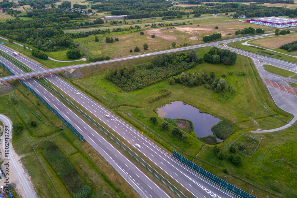 Aerial view of a highway intersection surrounded by green fields and trees. Cars are visible on the road, as well as sound barriers. The image shows a peaceful, rural setting. Aerial view of a highway intersection surrounded by green fields and trees. Cars are visible on the road, as well as sound barriers. The image shows a peaceful, rural setting.