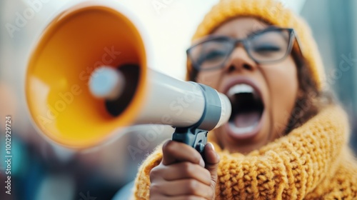 A woman in a yellow scarf and hat passionately shouts through a yellow megaphone on a bustling street, capturing the vibrancy and energy of public activism and protest. photo