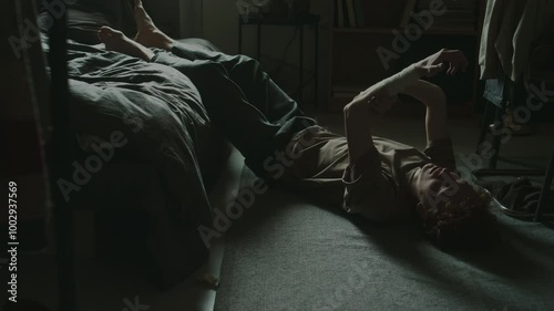Devastated young man lying on floor of his room with legs thrown on messy bed looking at his wrist wrapped in bandage and touching arm