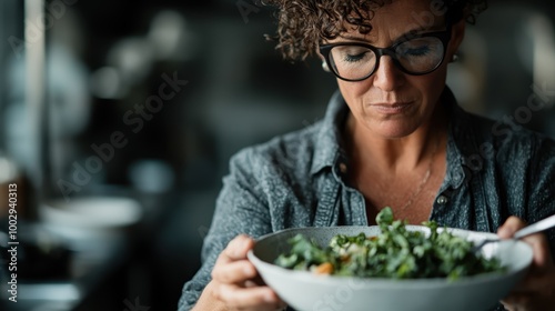 A woman with curly hair and glasses focuses solemnly on a bowl of salad, conveying a deep appreciation for healthy living and culinary artistry. photo