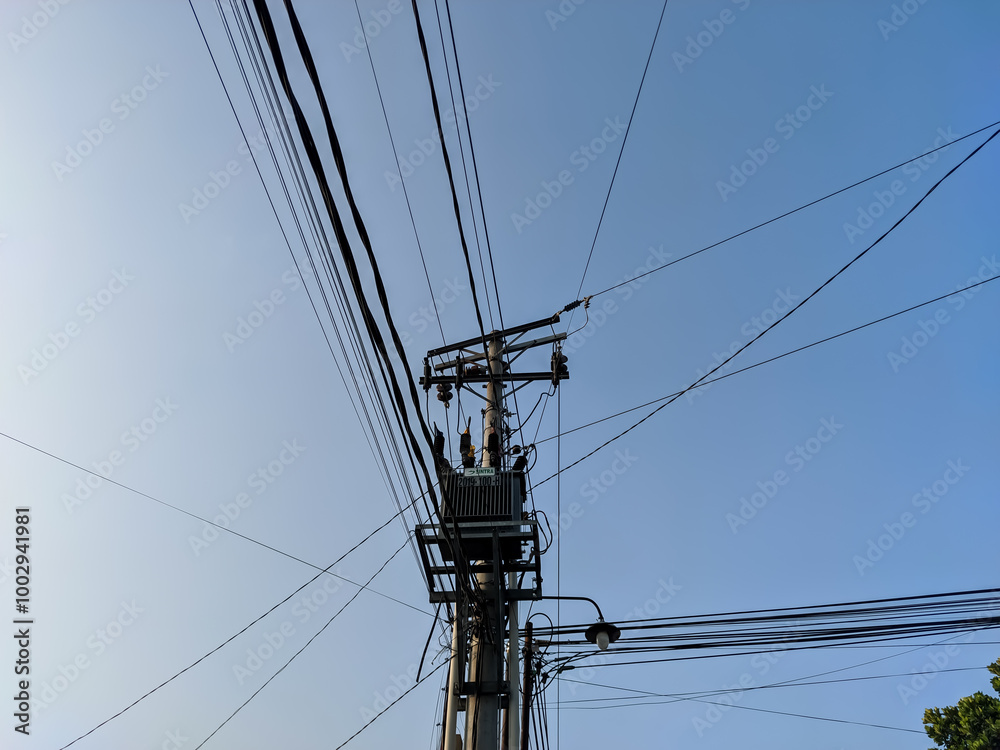 A tall utility pole stands against the backdrop of a clear blue sky, its frame crisscrossed by a complex web of cables. The intricate tangle of wires weaves in and out. A tall utility pole stands against the backdrop of a clear blue sky, its frame crisscrossed by a complex web of cables. The intricate tangle of wires weaves in and out.