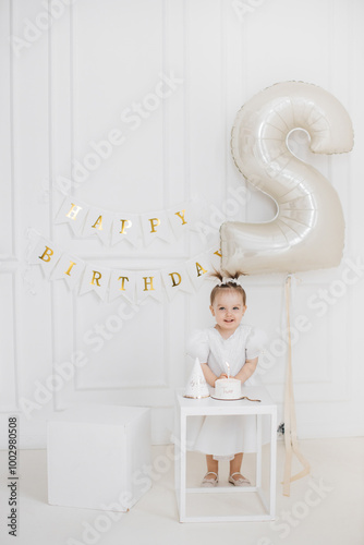 Smiling Caucasian toddler girl celebrating second birthday. White dress, festive hat, and cake with candle. Joyful expression, standing next to large number two balloon in bright room. photo