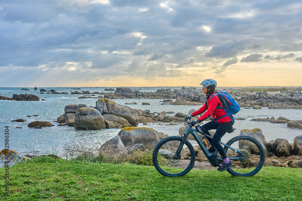 Obraz premium nice senior woman cycling with her electric mountain bike at the rocky rose granite coast at Meneham site in Brittany, France next to Kerouarn