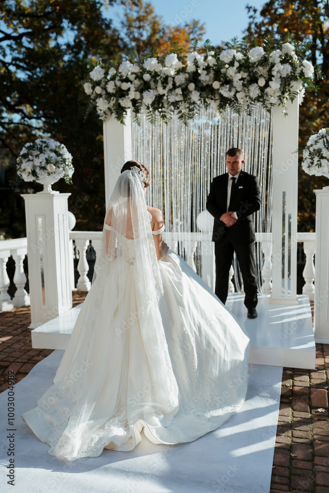 Fototapeta premium A bride and groom stand in front of a white archway. The bride is wearing a white dress and a veil, while the groom is wearing a black suit. The scene is set in a garden, with flowers