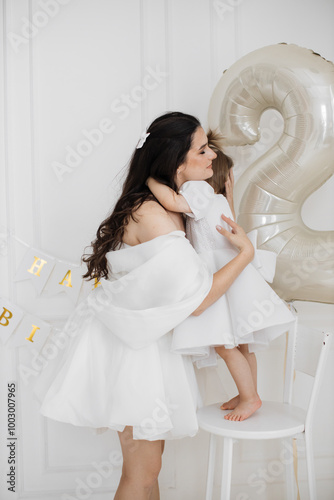 Young Caucasian mother and toddler daughter celebrating second birthday together. Mother kissing child, both wearing white dresses, large number two balloon in background. photo