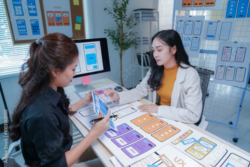 Two young ui/ux designers are discussing user interface design for a mobile application, holding a meeting at the office. They are working together to improve the user experience