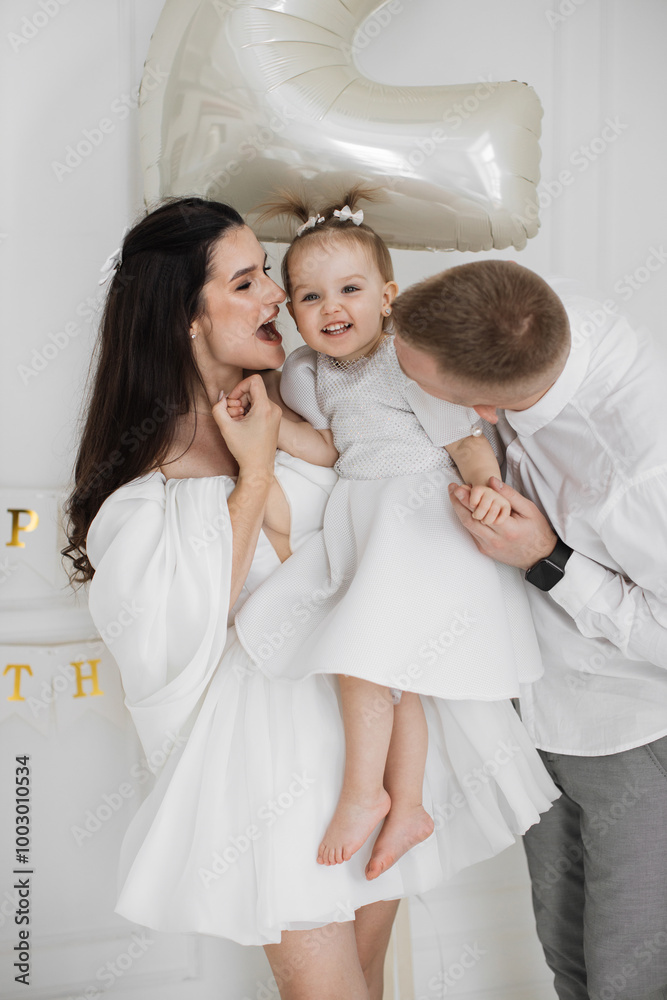 Caucasian parents with young daughter celebrating her birthday in bright room. Woman holding toddler while man playfully entertains. Caucasian parents with young daughter celebrating her birthday in bright room. Woman holding toddler while man playfully entertains.