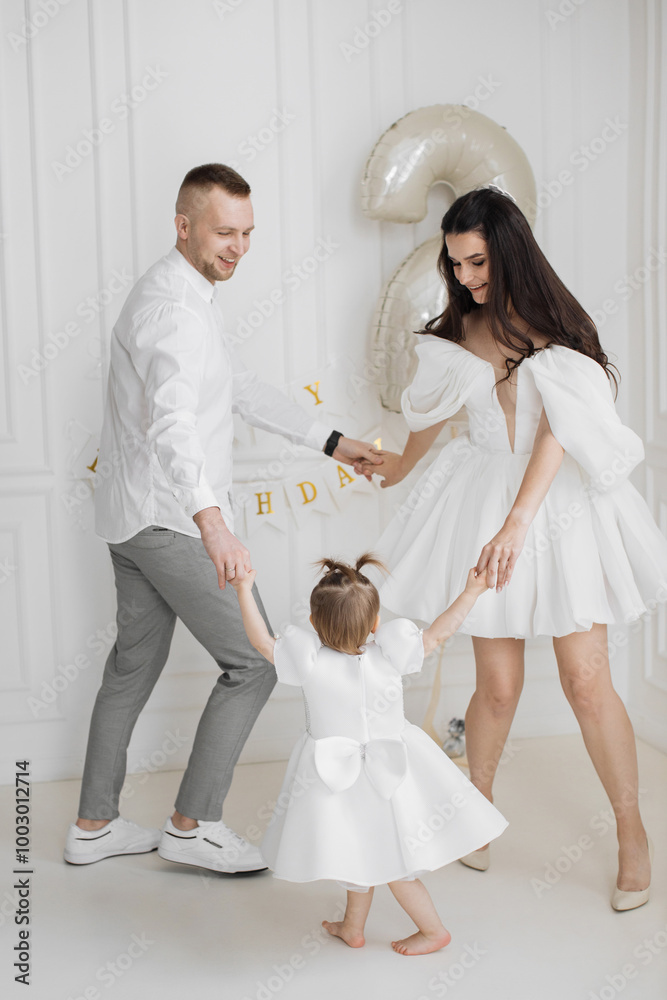Obraz premium Caucasian family celebrating child's birthday. Parents dancing with young daughter in white room decorated with happy birthday banner. Father mother and child dressed elegantly sharing joyful moment