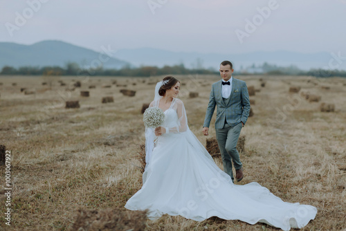 A bride and groom are walking through a field of hay. The bride is wearing a white dress and the groom is wearing a suit. The scene is peaceful and romantic