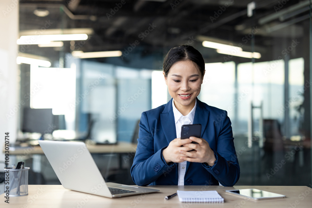 Asian businesswoman in blue suit using smartphone at office desk with laptop and notepad. Smiling, focused on communication in professional work environment. Concept of business technology Asian businesswoman in blue suit using smartphone at office desk with laptop and notepad. Smiling, focused on communication in professional work environment. Concept of business technology