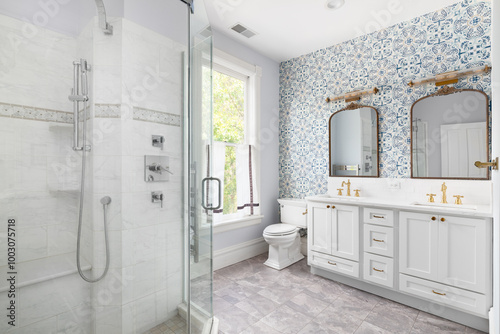A bathroom with a blue wallpaper accent wall, gold mirror and faucets, grey cabinet, tiled flooring, and a walk-in shower with a chrome showerhead.