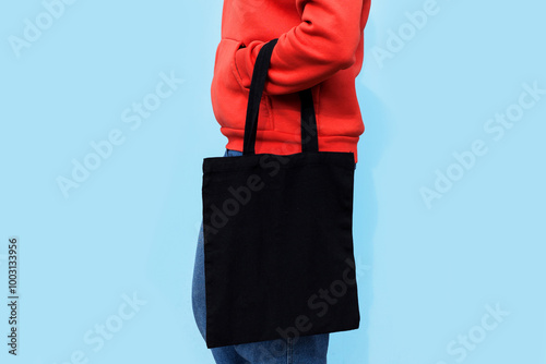 Woman in red hoodie holding tote canvas blank eco bag on street light blue minimal wall background. Female consumer hold black textile shopper. template or place for your design, text, logo photo