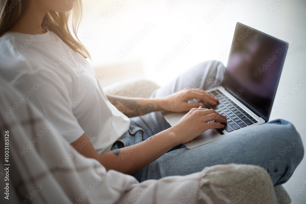 Naklejka premium Close up image of woman hands while she sits comfortably in a cozy chair, working on her laptop in a bright, sunlit room. She appears focused and relaxed, embodying a modern, remote work lifestyle.