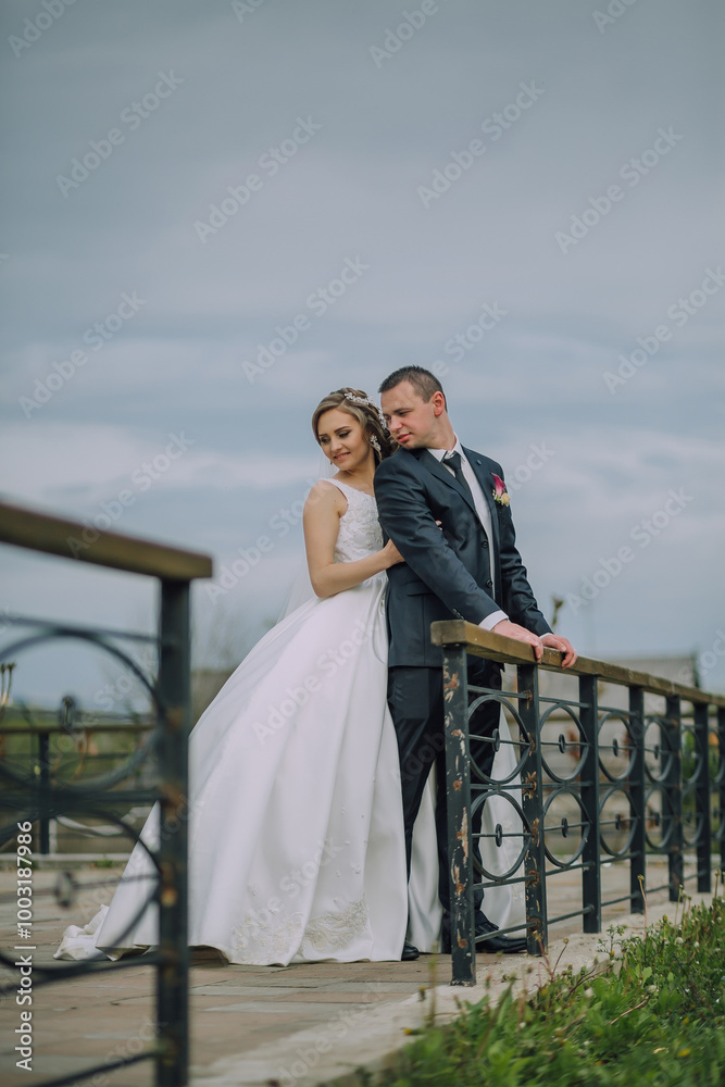 A bride and groom are standing on a bridge, posing for a picture. The bride is wearing a white dress and the groom is wearing a suit. Scene is romantic and happy A bride and groom are standing on a bridge, posing for a picture. The bride is wearing a white dress and the groom is wearing a suit. Scene is romantic and happy