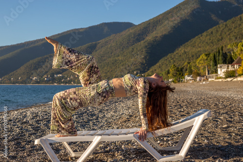 A girl in trousers and a top is standing on a chaise longue in the bridge. Acrobatics on the beach with mountain and sea views