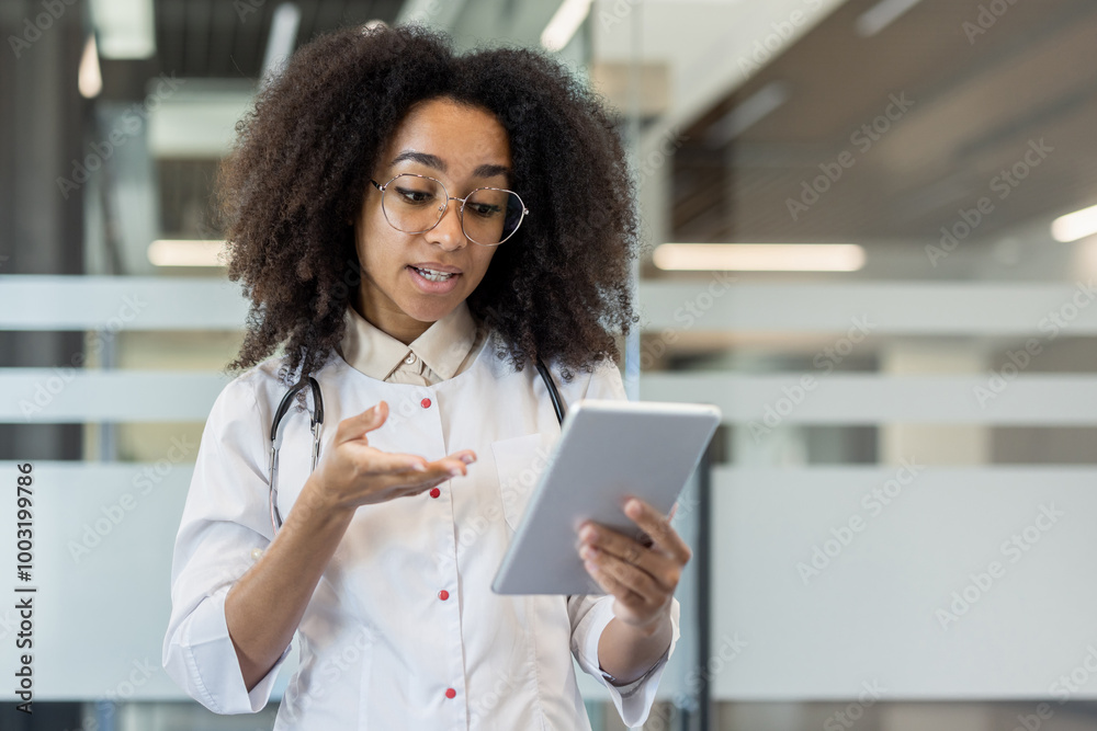 African american young female doctor standing in hospital in white uniform talking and consulting on video call on tablet