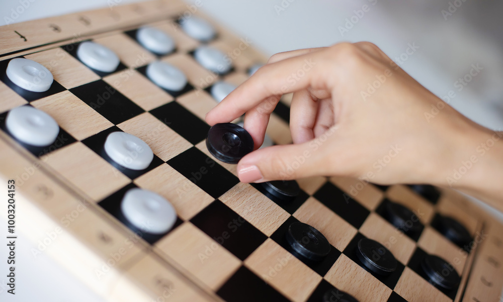 A close-up of a person’s hand moving a black checker piece on a wooden board. The image captures focus and strategy during a checkers game, emphasizing concentration and thoughtful gameplay. A close-up of a person’s hand moving a black checker piece on a wooden board. The image captures focus and strategy during a checkers game, emphasizing concentration and thoughtful gameplay.