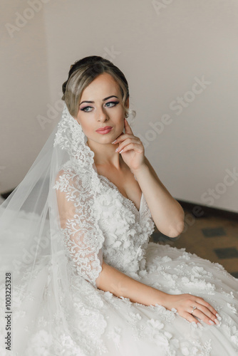 A woman in a white wedding dress sits on the floor with a lace veil on her head. She is wearing a ring on her finger and has her hand on her chin. Concept of elegance and sophistication