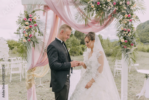 A bride and groom are getting married under a pink arch. The bride is wearing a white dress and the groom is wearing a black suit. They are standing under a pink canopy