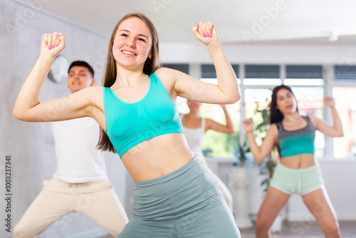 Female teen performs choreographic exercises and teaches energetic mobile social dance vogue together with friends. Young girls and guy repeat movements, train in spacious studio photo