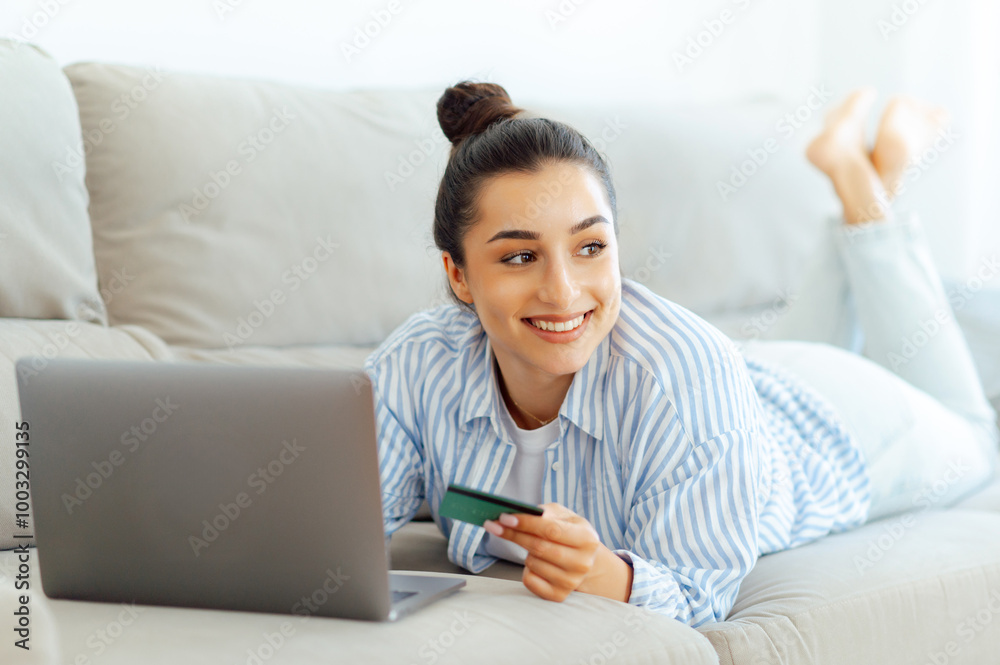 Charming happy caucasian woman in casual clothes lies on a couch in a home living room with a laptop, holding a credit card, making shopping or online banking in mobile app, looks away and smiles Charming happy caucasian woman in casual clothes lies on a couch in a home living room with a laptop, holding a credit card, making shopping or online banking in mobile app, looks away and smiles