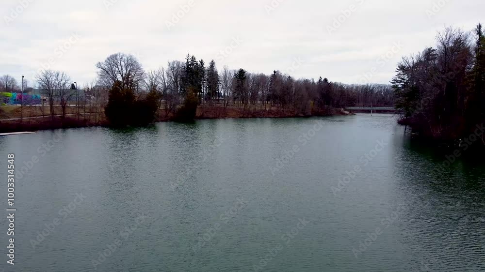 Drone moving quickly toward a small bridge over Lake Lisgar in Tillsonburg, Ontario. Camera passes by a small dock with a lovely background of leafless trees on a slightly overcast day. Drone moving quickly toward a small bridge over Lake Lisgar in Tillsonburg, Ontario. Camera passes by a small dock with a lovely background of leafless trees on a slightly overcast day.