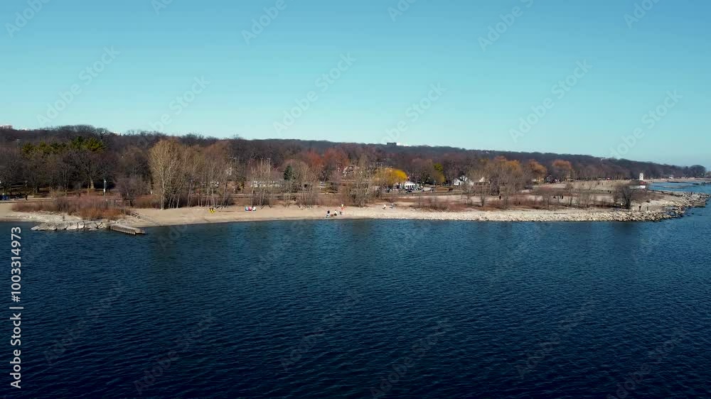 Drone orbit shot of Lake Ontario's coast on a lovely sunny day. People enjoy playing by the shore while a bird flies above them. We see a forest in the background with leafless trees. Drone orbit shot of Lake Ontario's coast on a lovely sunny day. People enjoy playing by the shore while a bird flies above them. We see a forest in the background with leafless trees.