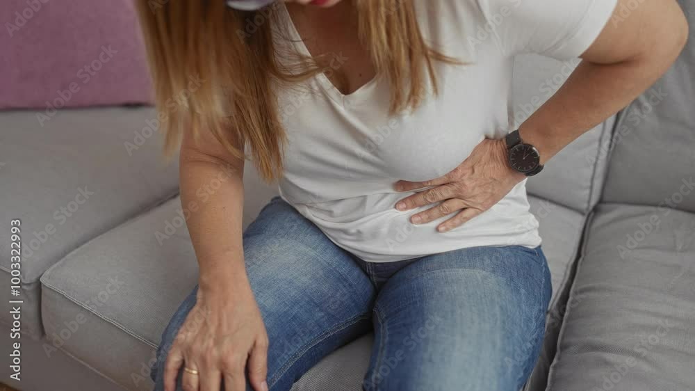 Woman experiencing stomach pain while sitting on a grey sofa at home in a casual white shirt and blue jeans Woman experiencing stomach pain while sitting on a grey sofa at home in a casual white shirt and blue jeans
