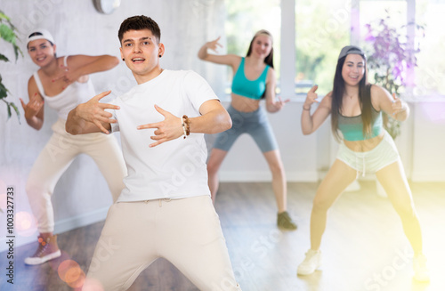 Positive smiling male teenager learns social dance, perform movements in choreographic class in company of peers. Young girls in ball cap and guy repeat movements, train in spacious training hall. photo