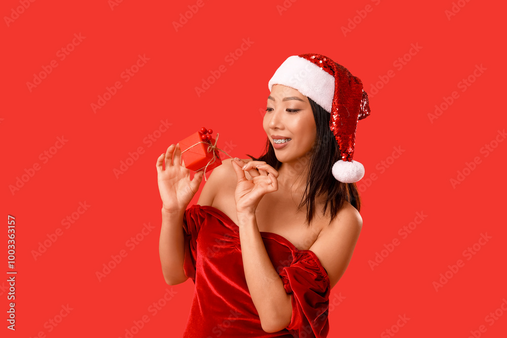 Young Asian woman in Santa hat with Christmas gift box on red background