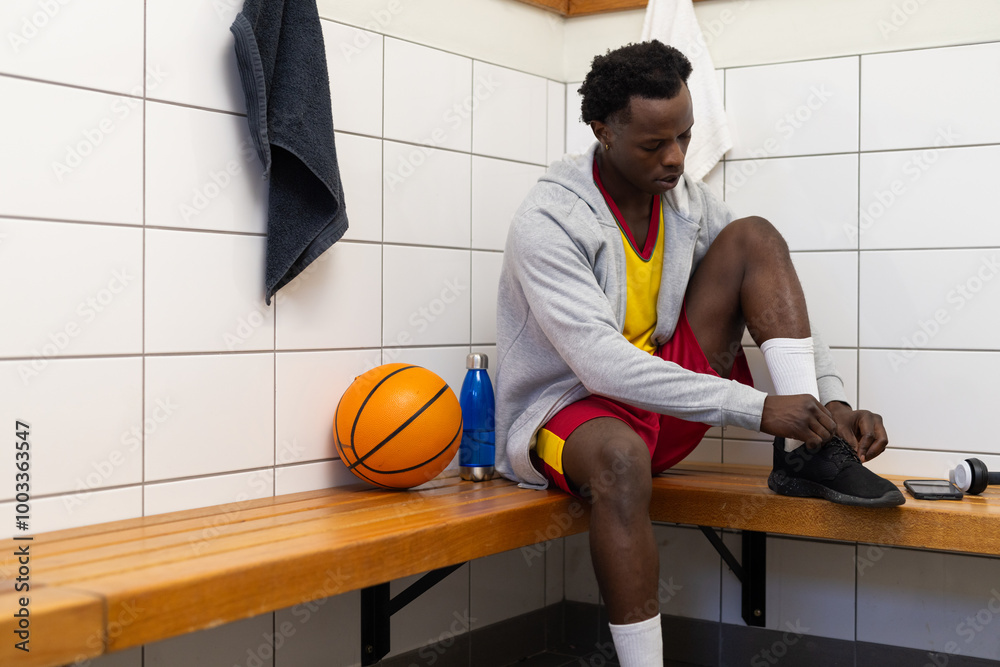 Tying shoelaces, basketball player sitting on bench in locker room with ball, copy space Tying shoelaces, basketball player sitting on bench in locker room with ball, copy space