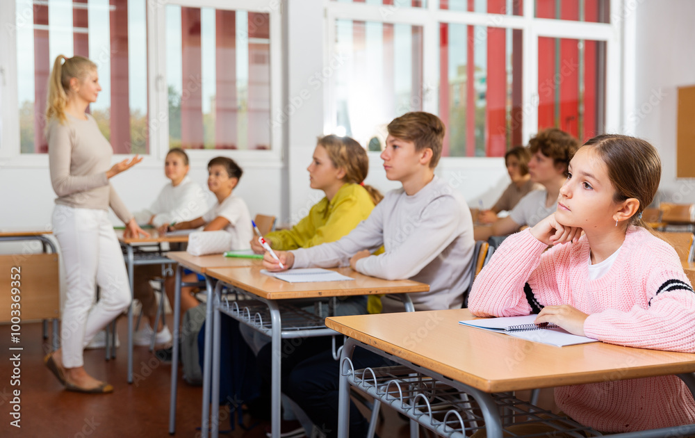 Fototapeta premium Focused teenage students studying in classroom with teacher, writing lectures in workbooks