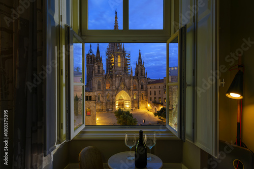 Canvas Print A bottle of wine and two wine glasses on a small table under an open window with the illuminated Barcelona Cathedral in view at night, in Barcelona, Spain
