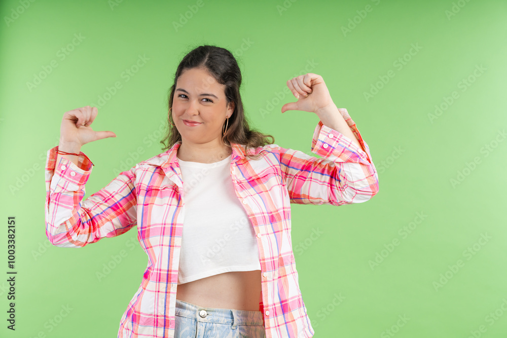 Young woman pointing her thumbs at herself with a proud expression on green background Young woman pointing her thumbs at herself with a proud expression on green background