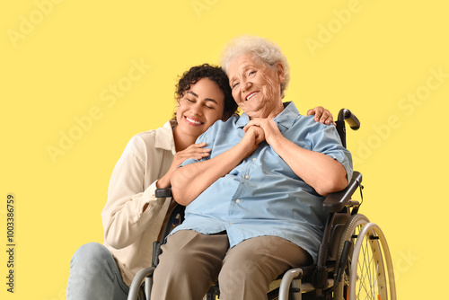 Young African-American female medical worker with elderly woman on wheelchair against yellow background