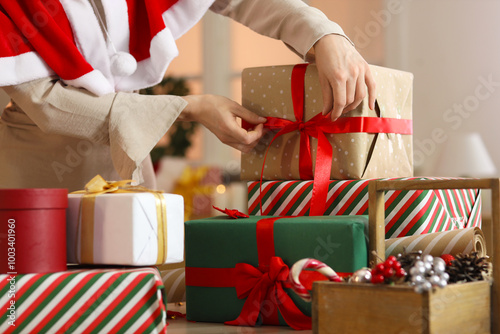 Beautiful young woman in Santa cape tying bow while wrapping Christmas gift boxes at home, closeup