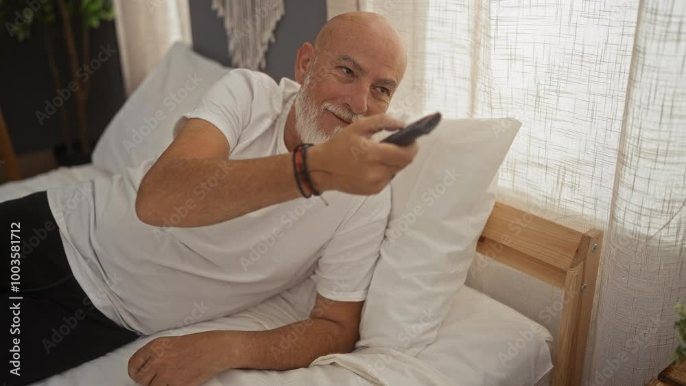 Man lying in bed in his bedroom at home holding a remote in a relaxed indoor setting, appearing happy and content with a grey beard under natural daylight. Man lying in bed in his bedroom at home holding a remote in a relaxed indoor setting, appearing happy and content with a grey beard under natural daylight.