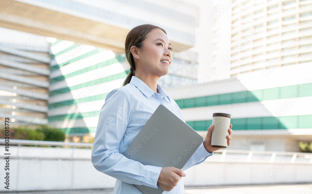 Asian woman stands outdoors, smiling while holding a laptop and a coffee cup. She is dressed in a collared shirt and appears relaxed in a modern urban setting on a clear day. Asian woman stands outdoors, smiling while holding a laptop and a coffee cup. She is dressed in a collared shirt and appears relaxed in a modern urban setting on a clear day.