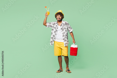 Billede på lærred Full body young Indian man he wearing beach shirt hat rest near hotel pool hold cooler fridge drink orange cocktail say cheers isolated on plain green background