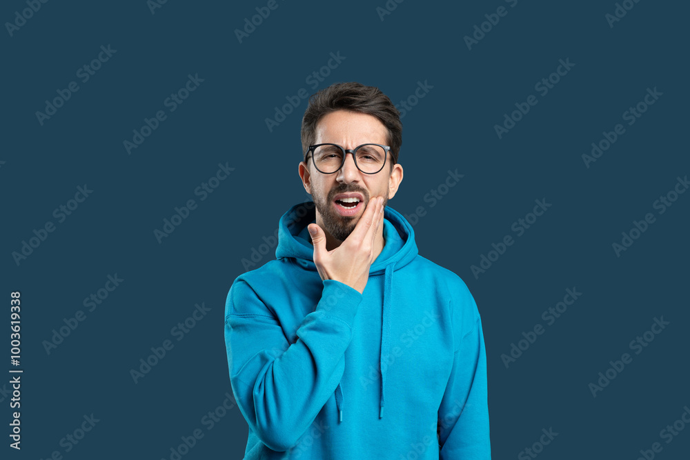 A man wearing a blue hoodie displays a pained expression while holding his chin. The dark background adds focus to his discomfort, possibly indicating a toothache or jaw pain. A man wearing a blue hoodie displays a pained expression while holding his chin. The dark background adds focus to his discomfort, possibly indicating a toothache or jaw pain.