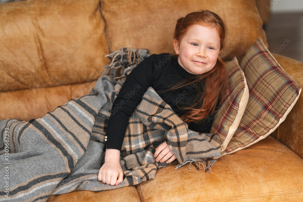 Fototapeta premium Cozy moments on a chilly afternoon with a young girl wrapped in a blanket on a comfortable couch at home