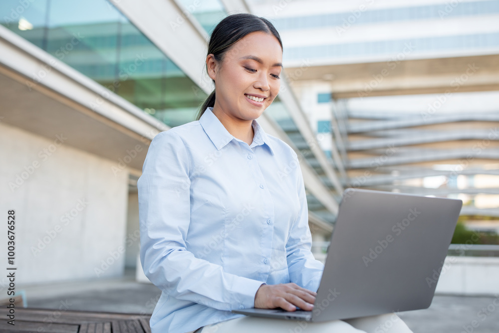 A young Asian woman smiles while working on her laptop outside a contemporary office building. She is focused and appears to enjoy her task in the pleasant environment. A young Asian woman smiles while working on her laptop outside a contemporary office building. She is focused and appears to enjoy her task in the pleasant environment.