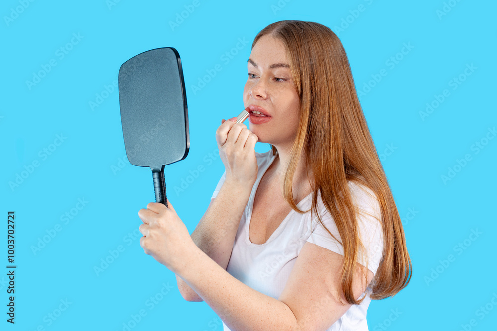 A young woman applies lipstick in front of a bright blue background, showcasing her makeup routine and confidence A young woman applies lipstick in front of a bright blue background, showcasing her makeup routine and confidence